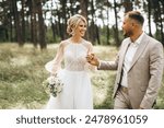 Wedding couple bride and groom walk hand in hand through a pine forest, sunlight filtering through the trees, highlighting their joyful expressions.