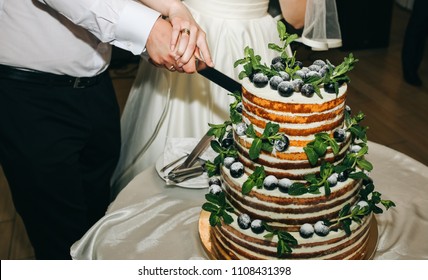 Wedding Couple Is Cutting Modern Rustic Cake. Open Sponge Dessert With Mint Leaves And Fresh Fruit Grapes On Top. Boho Style Wedding Cake. Groom In The Black Suit And Bride In White Elegant Dress.