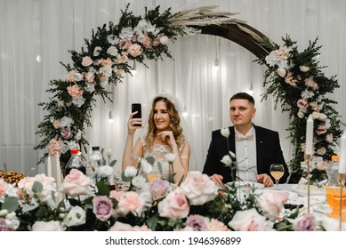 Wedding Couple Doing Silfi At The Wedding Table. Happy Lovers Are Photographed On The Phone During A Banquet. Wedding Feast. Holiday Food. Wedding Flowers And Decorations