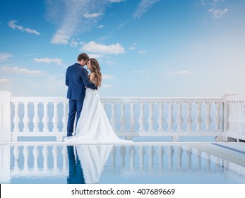 Wedding Couple Hugging And Standing With Her Back Outdoors Near The Pool. Happy Bride And Groom In Love On Their Honeymoon. Woman In An Elegant Dress A Man In The Suit. 