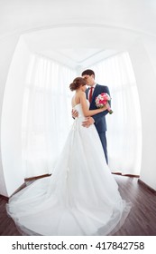 Wedding Couple In Love. Beautiful Bride In White Dress With Brides Bouquet And Handsome Groom In Blue Suite Standing And Embracing Each Other Indoors At Home, White Bright Room With Big Window High