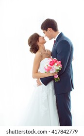 Wedding Couple In Love. Beautiful Bride In White Dress With Brides Bouquet And Handsome Groom In Blue Suite Standing And Embracing Each Other Indoors At Home, White Bright Isolated Background