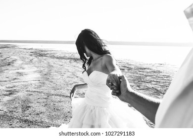 Wedding couple on sea beach. Sunny summer photo. Bride with hair down in off shoulder dress with train holds groom's hand. Ocean romantic. Seaside love story. Sand, water and horizon. Black and white.