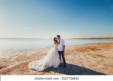 Wedding couple on the sea beach. Sunny summer photo. Bride with hair down in off shoulder dress with train. Ocean romantic ceremony. Seaside love story. Sand, water and horizon. Honeymoon photoshoot.