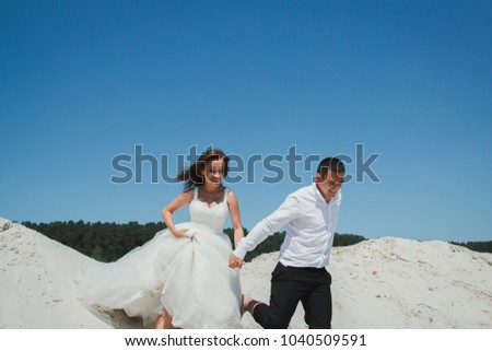 Wedding couple is running and jumping on the sandy lake beach. Funny laughing bride and groom in summer heat love story. Heating sun and weather. Travel leisure lifestyle true emotional photo.