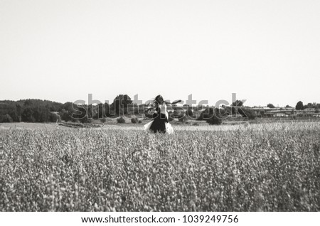 Wedding couple is spinning in the summer wheat field. Bride in white puffy dress hold groom's hands in black suit. Green trees and houses on the horizon. Summer heat and love story. Black and white.