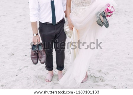Wedding couple is standing on the sand beach barefoot and holding their shooes in hands.