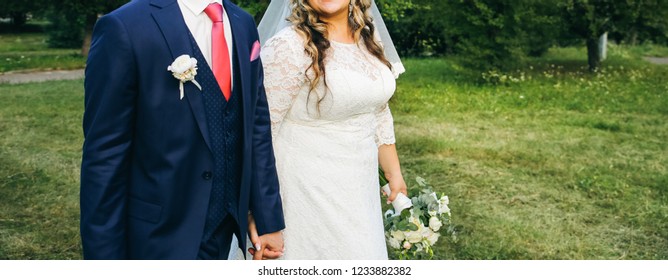 Wedding Couple Walking In The Green Park. Curvy Bride In White Lace Dress And Groom Are Holding Hands. Overweight Happy People. Love Story Outdoors. Beautiful Bouquet.