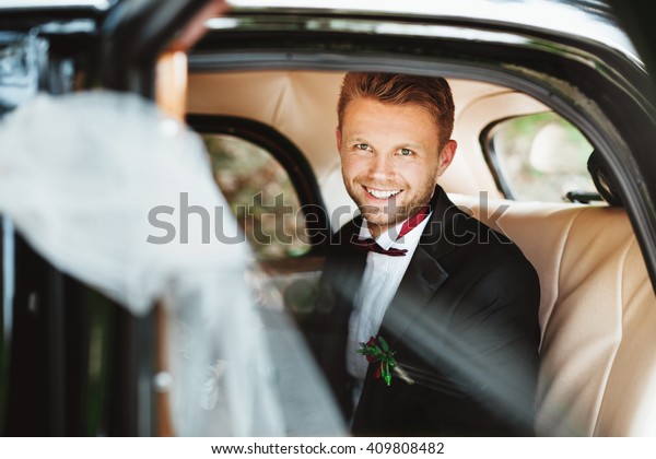 Wedding day. Bridegroom sitting in wedding car,\
looking at camera and smiling. Decorations. Sunny day. Waist up,\
closeup