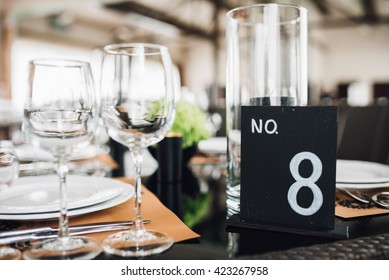 Wedding Decor. Reception. Table Number 8 On Black Wooden Stand. Glasses And Cutlery On Side. Steampunk Style