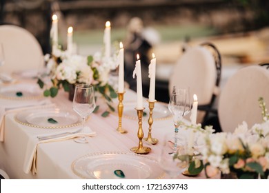 Wedding dinner table at reception. Beautiful white delicate candles burn in metal candlesticks, against background of white and pink tablecloth, flower arrangements and glass plates with gold beads