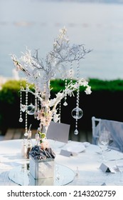 Wedding dinner table reception. Wedding table decoration - white branch from a tree, crystal pendants, candles in glass spheres, on a white table with a mirror, against the sea