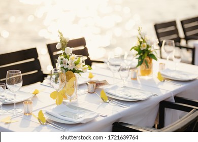 Wedding dinner table reception. Rectangular tables with white tablecloth, floral arrangements lemons in vases. Yellow paper butterflies are scattered on table. On beach, overlooking sunset at sea.