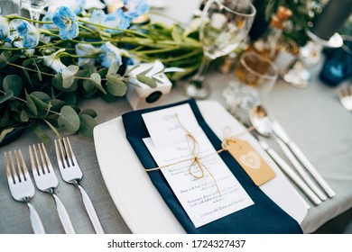 Wedding dinner table reception. A square plate with a blue cloth towel, knives and forks next to the plate. Flower composition with eucalyptus leaves in the center of the table and burning candles.