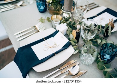 Wedding dinner table reception. A square plate with a blue cloth towel, knives and forks next to the plate. Flower composition with eucalyptus leaves in the center of the table and burning candles.