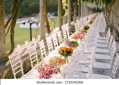 Wedding Dinner Table Reception. A Very Long Wedding Table Going Into Perspective. Bouquets Of Yellow, Orange, Pink Roses On Tables. Glass Chairs Chiavari, Tiffany Made Of Transparent Plastic