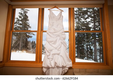 Wedding Dress Hanging In A Window Overlooking Pine Trees And A Lake During Winter.