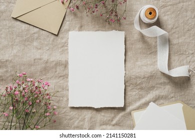 Wedding flat lay composition with blank paper card mockup, silk ribbon, envelopes, gypsophila flowers on linen tablecloth. Wedding invitation card design