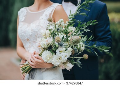 Wedding. The Groom In A Suit And The Bride In A White Dress Standing Side By Side And Are Holding Bouquets Of White Flowers And Greenery