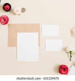 Wedding invitation cards, craft envelope, pink and red rose flower buds and white carnation on pale pastel beige background. Workspace with paper blank. Flat lay, top view - Shutterstock ID 651159475