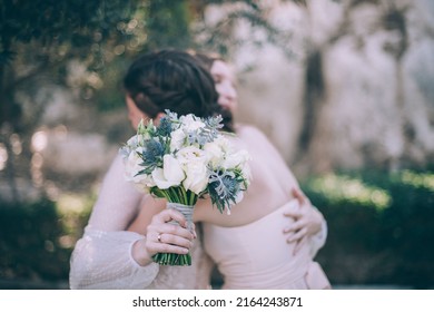 Wedding lgbt ceremony outdoors. Two brides in white wedding dresses gently hug each other. Bouquet and bride's hand with wedding ring in focus in the foreground