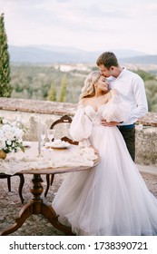 Wedding At An Old Winery Villa In Tuscany, Italy. A Wedding Couple Is Standing Near The Table For A Wedding Dinner, The Groom Hugs The Bride By The Waist.