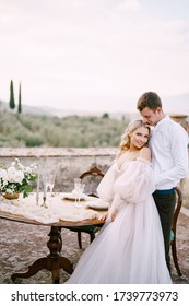 Wedding At An Old Winery Villa In Tuscany, Italy. A Wedding Couple Is Standing Near The Table For A Wedding Dinner, The Groom Hugs The Bride By The Waist.