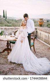 Wedding At An Old Winery Villa In Tuscany, Italy. A Wedding Couple Is Standing Near The Table For A Wedding Dinner, The Groom Hugs The Bride By The Waist.