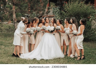 Wedding photo in nature. A brunette bride in a long white dress and her friends in nude dresses stand against a background of trees, smiling and holding their gypsophila bouquets. Young women.