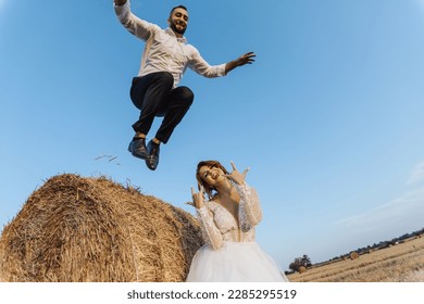 Wedding photo. A stylish groom in a white shirt jumps down from a hay bale, the bride is standing below. Bearded man. Red-haired bride. Style. Emotions - Powered by Shutterstock