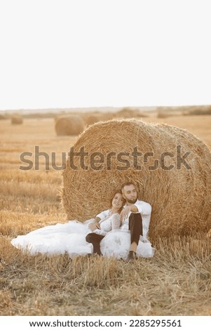 Wedding portrait. The bride and groom are sitting in an embrace on the ground, near a bale of hay. Red-haired bride in a long dress. Stylish groom. Summer. A sincere smile. On the background of hay