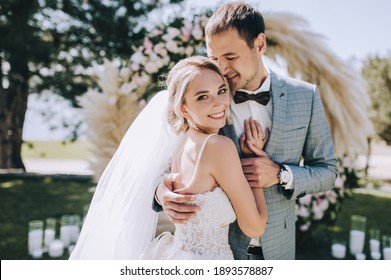 Wedding portrait of smiling newlyweds. Stylish groom in a gray checkered suit and a cute blonde bride in a white lace dress are tenderly hugging in the forest against the background of the arch.