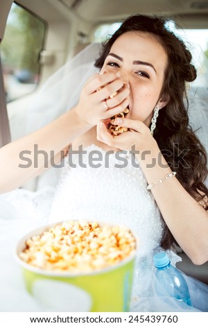 Wedding portrait of young bride sitting in car straight, greedily eats popcorn before movie, hairstyle and bright makeup. Strong expression, shallow depth of field, focus on eyes and a bit on mouth.