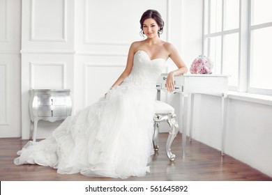 Wedding preparation. Beautiful young bride in white wedding dress indoors. Luxuty model sitting on chair with brides bouquet like at home in studio room with big window. Girl shows wedding fashion in - Powered by Shutterstock