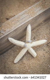 Wedding Rings Around Arm Of Starfish In Beach Sand Against Wooden Plank For Rustic Beach Wedding In Portrait Photo