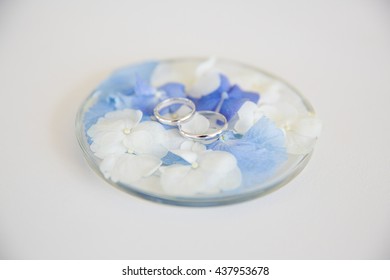 Wedding rings lie on tiny white and blue flowers on a glass plate - Powered by Shutterstock