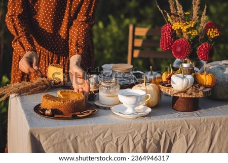 Wedding or romantic date decoration in the field with red dahlia. Beautiful happy young woman on a meadow arranging table for outdoor autumn event, tea party with homemade pumpkin cake. 