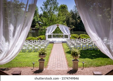 Wedding Venue Next To A Pond And Forest With White Chairs And A White Gazebo And A Brick Isle. 