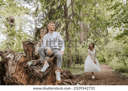 Wedding walk in the forest. The bridegroom is seated on a felled old tree in the foreground. Country wedding concept.