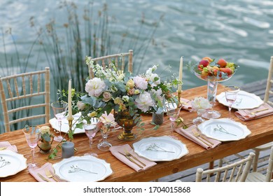 Wedding wooden table is standing on the pond shore, the water blurred background. Solemn table is set with plates, candles, bowl of fruits and beautiful bouquet of light pink chrysantemums and roses - Powered by Shutterstock