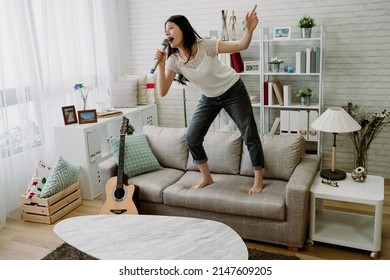 Weekend Day Asian Girl Is Standing On Couch Blowing Off Steam By Singing Karaoke At Home. Chinese Woman Holding Microphone With Exaggerated Body Movement Is Having Happy And Crazy Time Alone.