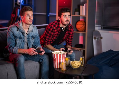 Weekend Entertainment. Involved Trendy Cute Guys Are Sitting On Couch And Holding Joystick While Playing Home Video Games. They Are Looking At Screen Absorbedly. Copy Space In The Right Side