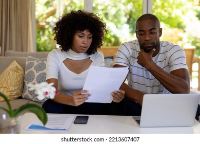 Weekend Fun At Home Together. Front View Close Up Of A Mixed Race Couple Sitting In The Living Room On The Couch, Looking At Paperwork And Using A Laptop Computer Together And Looking At Paperwork