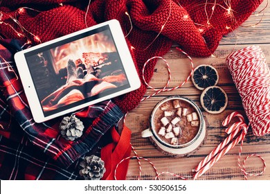 Weekend homely scene. Warm knit sweater and cup of hot cocoa with marshmallows. Christmas lollipop, led lights string and other holiday decor. Tablet pc with film, watching winter movies.