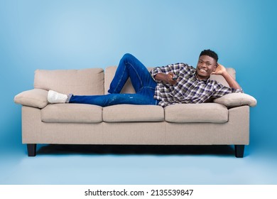 Weekend Leisure Activities. Millennial Black Man With Remote Control Switching Channels On TV While Lying On Couch Over Blue Background. African American Guy Watching Interesting Program On Television