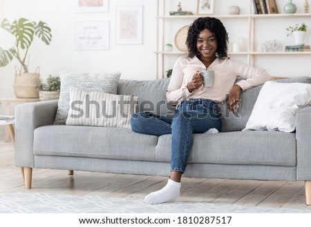 Weekend Leisure. Positive Black Woman Drinking Tea And Relaxing On Sofa At Home, Holding Cup With Hot Drink And Smiling At Camera, Copy Space
