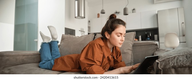 Weekend and lifestyle concept. Young woman lying on couch with digital tablet, scrolling social media, reading e-book or watching tv series on app. - Powered by Shutterstock