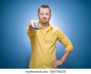 Weekend Time. Man Holding Remote Control And Watching TV - Movies Over Blue Background, Dresses In Yellow Shirt. Front View Of Guy Shows Television. Studio Shot