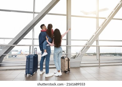 Weekend Travel. Family Of Three Standing Near Window In Airport And Pointing Away, Rear View Of Young Parents And Their Little Daughter With Suitcases Waiting For Flight At Terminal, Copy Space