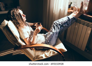 Weekends Finally. Woman In Blue Jeans Relaxing With Cup Of Tea In Armchair At Home, Daydreaming. Girl Enjoying Life, Bright Sunlight And Heat From Radiator. Cozy Home. Heating Season. Mortgage Concept